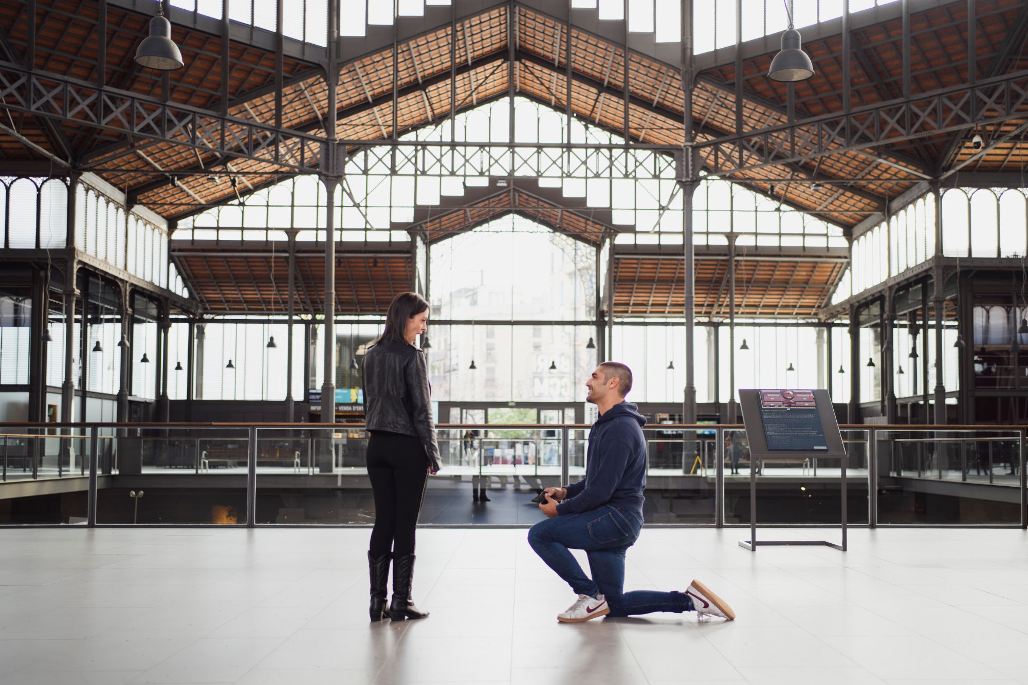Proposal at the Mercat del Born in Barcelona — kneeling partner presents the ring under the market's modernist iron-and-wood architecture